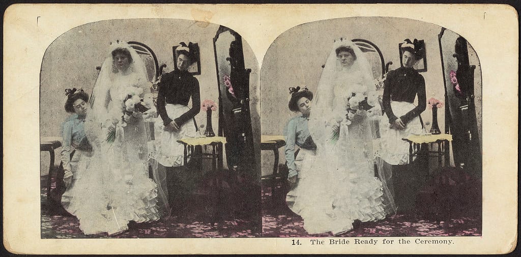 A stereograph from around 1900 showing a bride getting ready. A maid and seamstress help the bride dress while she poses, exp