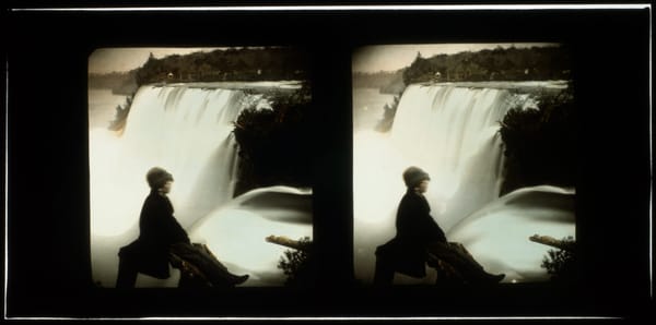 A grayscale stereograph slide of a person sitting in front of a large waterfall