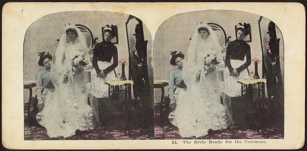 A stereograph from around 1900 showing a bride getting ready. A maid and seamstress help the bride dress while she poses, exp